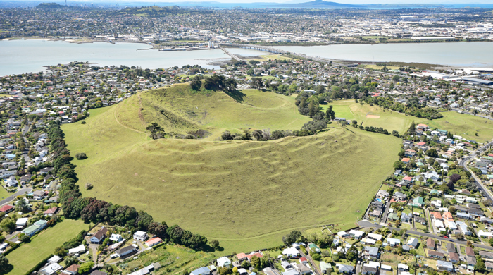 An aerial view of the mostly grassed maunga surrounded by houses. Māngere Bridge and the Manukau Harbour can be seen at the top of the image.