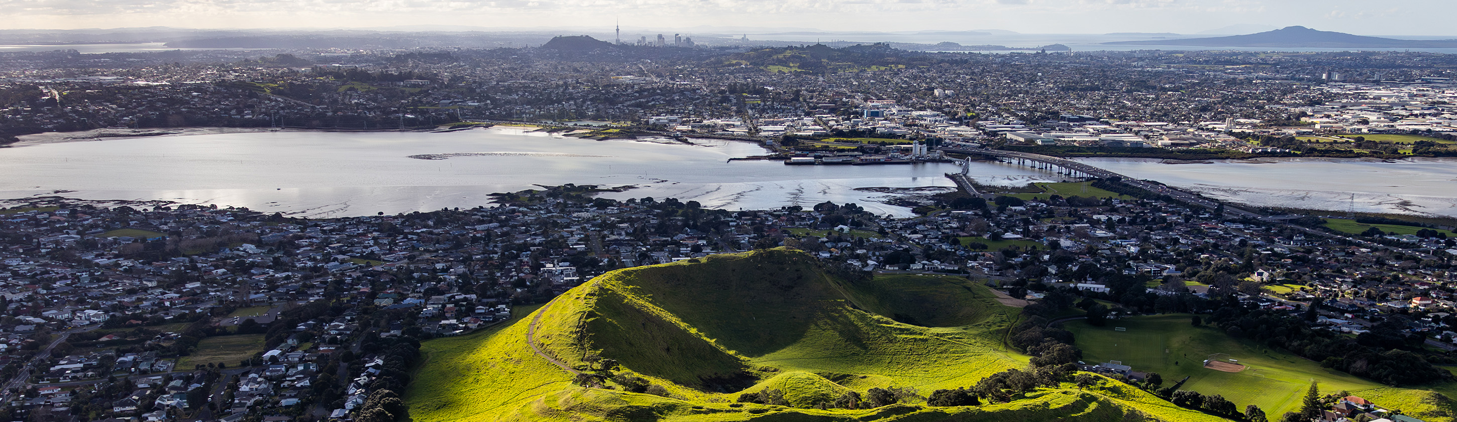 Aerial view of Māngere Mountain in South Auckland. Vibrant green grass and some trees cover the crater - an ancient and prominent cultural feature. The Māngere Inlet, part of the Manukau Harbour, Rangitoto Island and Mangere Bridge are to the north. To the right of the bridge is the historical portage where waka were carried over land between the Tamaki River and the Manukau Harbour. The sprawling urban landscape of Auckland city with its distinctive skyline and the Sky Tower is in the distance.