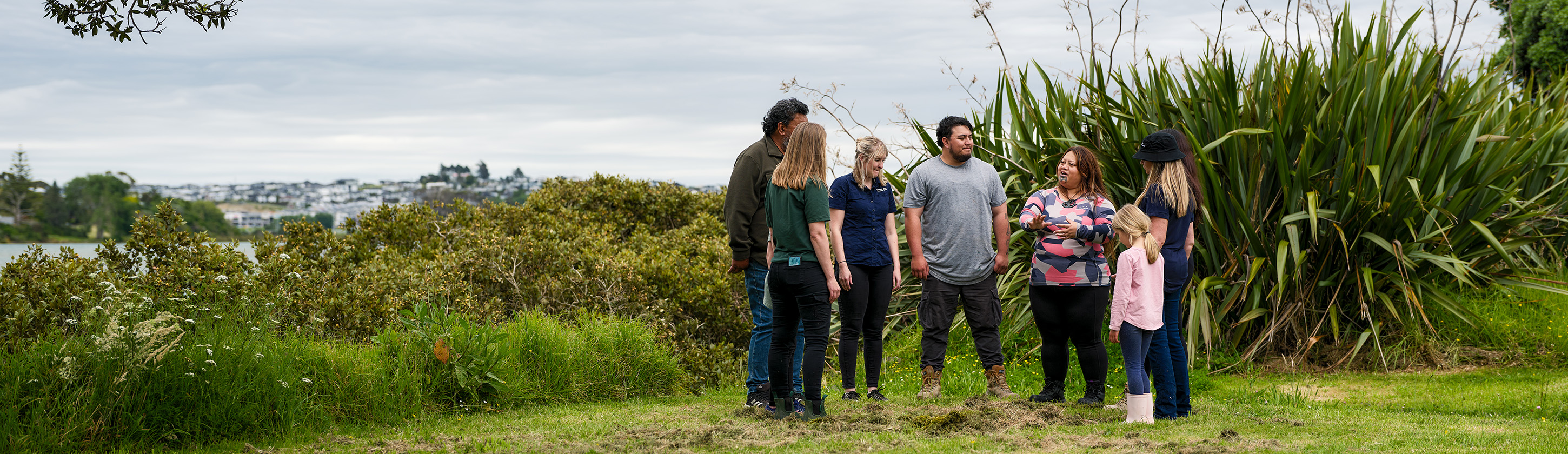 A group of people from iwi, community and Auckland Council are in a park in Ōrewa on Auckland’s nor-eastern shores. They are standing listening to the Māori woman in the group who is Te Ao Rosieur from Ngāti Manuhiri. A large flax/harakeke plant, lush greenery and mangroves are in the background. The image conveys how everyone can work together to support nature.