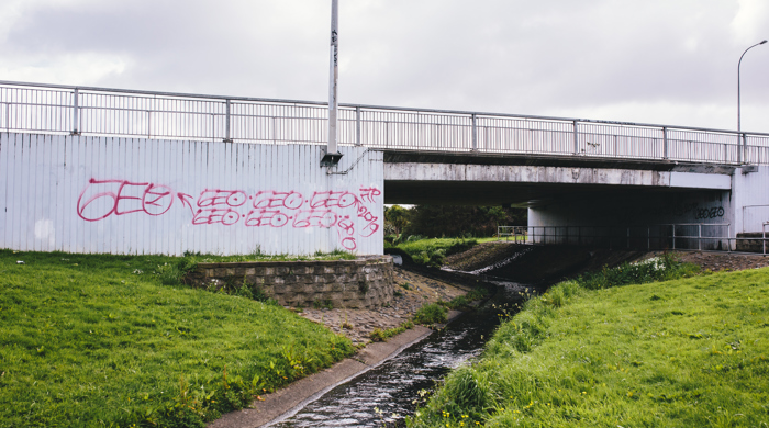 The stream flows under an urban bridge on a wide concrete channel with grass either side. Graffiti has been sprayed on the wall of the bridge.