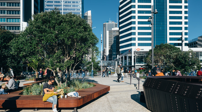 Seating and the outside of one of the mussel rope viewing areas are in the foreground with people, trees and high rise buildings in the background.