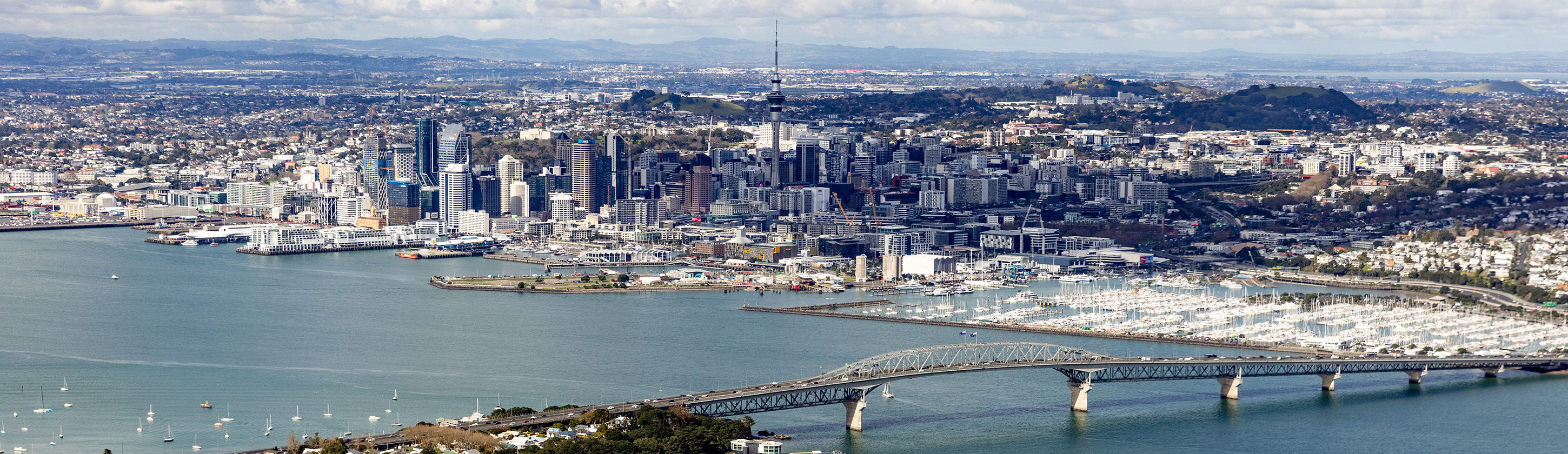 Aerial view of Auckland city looking south from Birkenhead, across the Waitematā. The iconic Sky Tower, high-rise buildings and sprawling urban areas sitting mostly on reclaimed land are in the middle of the picture. The Auckland Harbour Bridge connecting the city and the North Shore is in the foreground. Numerous boats are docked in the Westhaven Marina at the southern end of the bridge. Some volcanic cones, including the distinct dark green Maungawhau / Mount Eden are in the background.